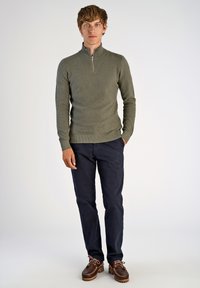 Olivgrüner Pullover mit Reißverschluss am Hals und strukturiertem Finish, kombiniert mit navyfarbenen Hosen und braunen Schnürschuhen mit kontrastierender Sohle.