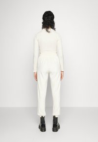 Kobieta z kręconymi, ciemnymi włosami stoi plecami, ubrana w dopasowany, długi rękaw kremowy top, pasujące spodnie i czarne buty sznurowane.