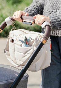 Szürke pulcsit viselő személy babakocsit tol, amelynek barna fogantyúja van, bézs pelenkázótáska van rákötve, és egy kis plüssjáték látható a táska zsebében.