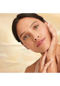 Ritratto di una persona con incarnato medio, occhi verdi e capelli scuri, che indossa un trucco minimale e una maglietta di colore chiaro, con le mani che toccano il viso.