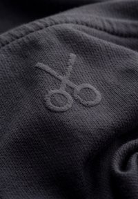 Donkergrijze stof met een reliëf schaaricoon. De textuur lijkt zacht en iets geweven, met gladde randen en subtiele details.