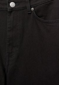 Jean en denim noir en gros plan montrant la poche avant, la boucle de ceinture et le bouton en métal avec les détails du nom de la marque.