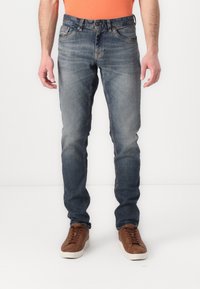 Jeans de mezclilla azul con corte ajustado, textura desgastada, diseño de cinco bolsillos y un lavado oscuro, combinados con zapatos marrones de cordones.