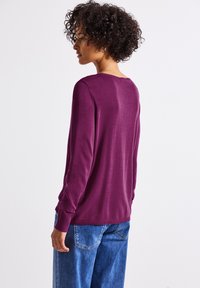 Lila langärmliger Pullover mit Rundhals und gerippten Bündchen, kombiniert mit hellblauen weit geschnittenen Jeans. Glatte Textur, minimale Details.