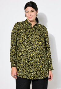 Camisa de botón con estampado de leopardo verde y acentos en negro y amarillo, con cuello y mangas largas, combinada con pantalones negros texturizados.