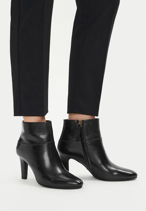 Czarne skórzane botki za kostkę z bocznymi zamkami, noszone z czarnymi obcisłymi spodniami na gładkim jasnym tle.
