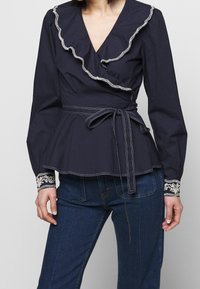 Mörkblå omfamnande blus har en volangkrage, långa ärmar med broderade manschetter och midjeband för en strukturerad silhuett.