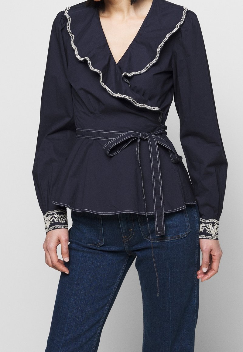 Mörkblå omfamnande blus har en volangkrage, långa ärmar med broderade manschetter och midjeband för en strukturerad silhuett.