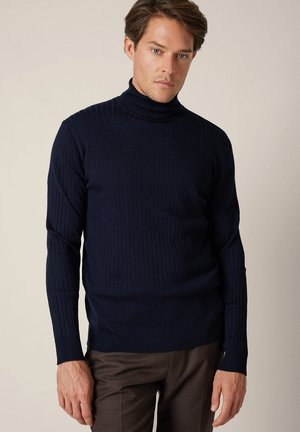 Męski model w granatowym prążkowanym golfie i brązowych spodniach, stojący na tle jednolitego beżowego tła.