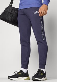 Jūras spēku jogger bikses ar baltu PUMA logo, ar sānu lentu dizainu, ribotām aprocēm un gludu tekstūru. Kopā ar melniem sporta apaviem.