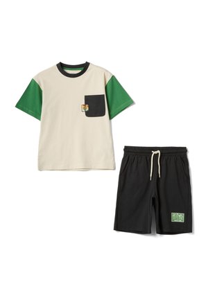 Beiges T-Shirt mit grünen Ärmeln und schwarzer Tasche mit kleinem Kaktus-Aufnäher, kombiniert mit schwarzen Shorts mit Kordelzug und grünem "Wild Life"-Aufnäher.