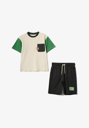Beiges T-Shirt mit grünen Ärmeln und schwarzer Tasche mit kleinem Kaktus-Aufnäher, kombiniert mit schwarzen Shorts mit Kordelzug und grünem "Wild Life"-Aufnäher.