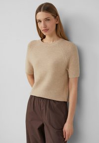 Beige Kurzarm-Strickpullover mit Rundhalsausschnitt und gerippten Akzenten, kombiniert mit lockeren braunen Hosen. Die Textur wirkt weich und fein.