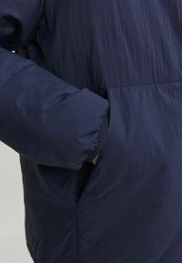 Tamno plava jakna s teksturiranom površinom. Ima pleteni manžet na rukavu i bočnu džep, naglašavajući pojednostavljen dizajn.