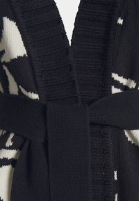 Gros plan d'un cardigan noir en tricot avec col côtelé et ceinture, avec des motifs abstraits blancs sur les côtés.