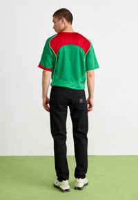 Vīrietis stāv uz zaļā grīdas, ģērbies zaļā un sarkanā īsajā piedurknē apģērbtā krekla, melnās biksēs un baltos sporta apavos, pagriezies prom pret baltu fona.