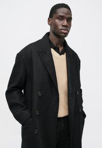 Cappotto doppio petto nero in misto lana con rever a lancia, caratterizzato da due bottoni neri e tasche, indossato sopra un maglione beige lavorato a maglia.