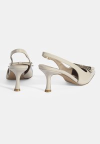 Talons compensés en cuir verni beige avec un détail en nœud, talons fins et design à bout rond. Texture lisse et finition brillante subtile.