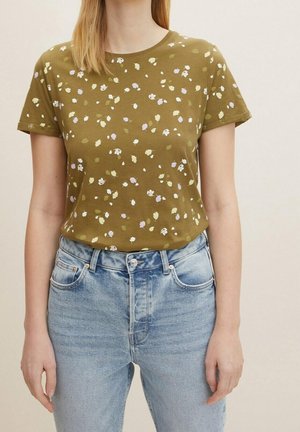 Frau trägt ein olivgrünes T-Shirt mit kleinem weißem und helllila Blumenmuster und hellblaue, hoch taillierte Jeans.