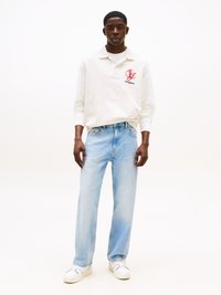 Polo a maniche lunghe beige chiaro con un logo rosso, abbinato a jeans azzurri larghi e sneakers bianche. Design semplice e casual.