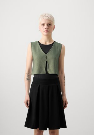 Zöld kötött, rövidített, V nyakú mellény, elől gombbal. Fekete, testhez simuló, ujjatlan felső alatta és fekete, pliszírozott szoknya.