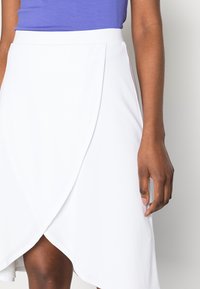 Biała asymetryczna spódnica wykonana z gładkiego materiału, z projektem w stylu kopertowym. Częściowo widoczna ręka oparta na talii.