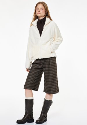 Joven mujer lleva chaqueta de forro polar blanca, pantalones cortos marrones de cuadros hasta la rodilla, botas negras hasta la mitad de la pantorrilla y calcetines blancos, de pie con una mano en el bolsillo.