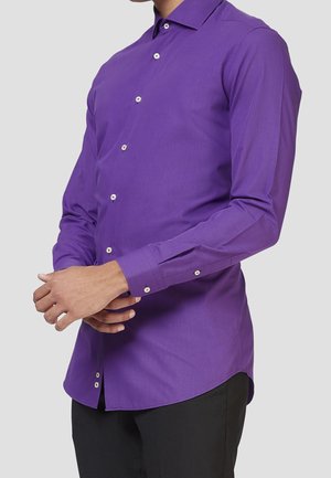 Homme portant une chemise ajustée à manches longues violette avec des boutons blancs, debout devant un fond clair uni.