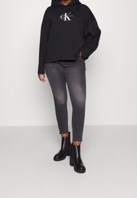 Felpa nera con cappuccio e logo "CK", jeans skinny grigi e stivali neri alla caviglia con tacco chunky, su uno sfondo neutro.
