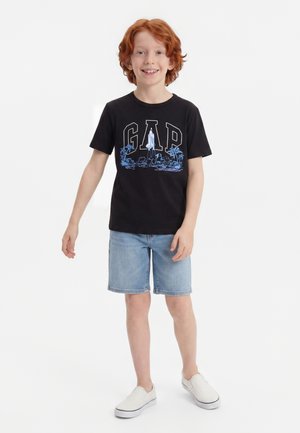 Garçon roux souriant portant un t-shirt noir GAP avec un motif bleu, un short en jean clair et des chaussures blanches à enfiler, debout sur un fond blanc.