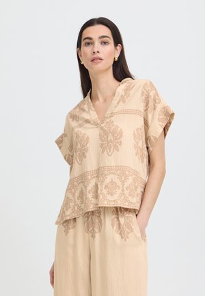 Kobieta o długich ciemnych włosach, ubrana w beżowy top z krótkim rękawem i dekoltem w serek oraz dopasowane spodnie w kwiatowy wzór, stojąca na jednolitym tle.