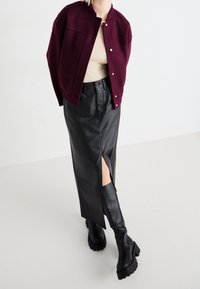 Veste en laine bordeaux avec boutons-pression, haut beige, jupe midi en cuir noir avec fente latérale, et bottes noires montantes épaisses.