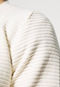 Nahaufnahme eines cremefarbenen, gerippten Strickpulloverärmels, der strukturierte horizontale Rippen und Nahtdetails im Schulterbereich zeigt.