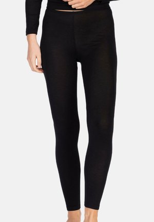 Frau trägt enge schwarze Leggings und ein passendes langärmeliges Oberteil, steht vor einem schlichten hellen Hintergrund.