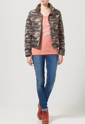 Chaqueta de camuflaje acolchada en tonos verdes y marrones con un cuello alto, combinada con una camiseta gráfica rosa claro y jeans azules.