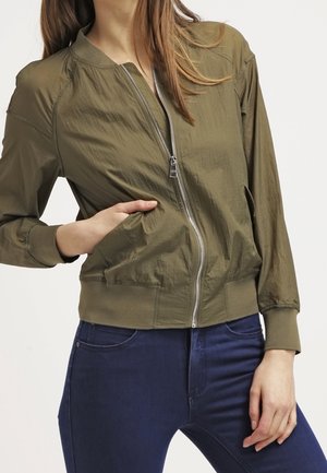 Kvinde iført en olivengrøn bomberjakke med lynlås, med den ene hånd i lommen og mørkeblå jeans mod en hvid baggrund.