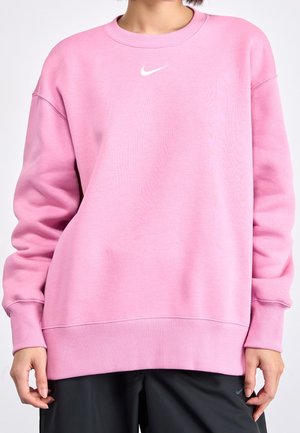 Persoon draagt een losse roze Nike-trui met geribbelde manchetten en zoom, gecombineerd met een zwarte broek, zichtbaar van hals tot heupen.