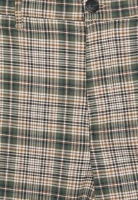 Närbild av brun, grön och svart rutigt tyg med en svart knapp och en del av en ficka synlig.