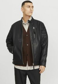 Svart läderjacka med quiltade axeldetaljer, silverfärgad dragkedja och ståkrage, lager på lager över en brun cardigan och en randig skjorta.