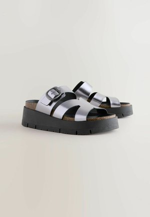 Metalliset hopeanväriset liukusandaalit korkkipohjalla, joissa on leveät hihnat ja musta kumipohja. Säädettävä solk yksityiskohta sivulla.