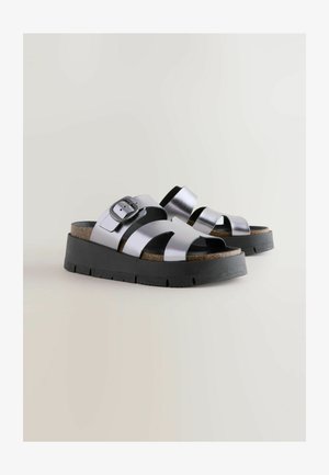 Metalliset hopeanväriset liukusandaalit korkkipohjalla, joissa on leveät hihnat ja musta kumipohja. Säädettävä solk yksityiskohta sivulla.