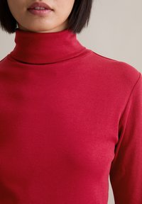 Red, ribbed turtleneck top featuring a close-fitting design and smooth texture. Notable high collar and minimalistic style.