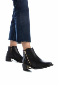 Botas de tobillo negras con cuero suave y acentos de textura de cocodrilo, con cremalleras doradas y un tacón bajo en bloque. Llevadas con denim recortado.