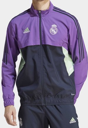 Mężczyzna w fioletowo-czarnej dresowej kurtce i spodniach Adidas z logo Realu Madryt, stojący z rękami swobodnie opuszczonymi po bokach.
