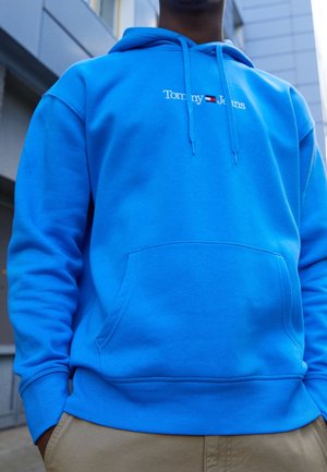 Person iført en lys blå Tommy Jeans hoodie med frontlomme og beige bukser, stående udendørs nær moderne bygninger.