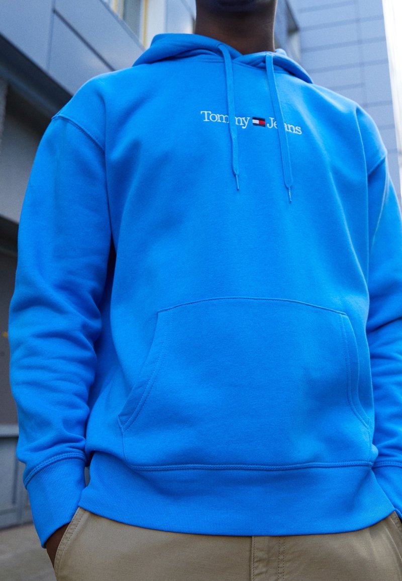 Personne portant un sweat à capuche Tommy Jeans bleu vif avec une poche avant et un pantalon beige, debout en plein air près de bâtiments modernes.