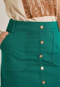 Jupe en denim teal avec taille haute, fermeture à boutons sur le devant et deux poches latérales. Les détails de couture rehaussent sa texture.