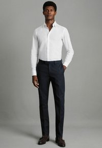 Bijela košulja dugih rukava u kombinaciji s tamnoplavim hlačama. Model nosi smeđe elegantne cipele. Glatka tkanina s krojem prilagođenim tijelu, minimalno ukrašena.