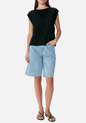 Femme portant un haut noir sans manches, un short en jean bleu clair longueur genou et des sandales noires à brides, debout avec une main dans la poche.