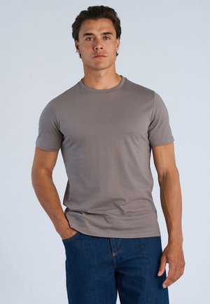 Man staat recht vooruit kijkend, draagt een effen grijs T-shirt met korte mouwen en een blauwe spijkerbroek met één hand in zijn zak.